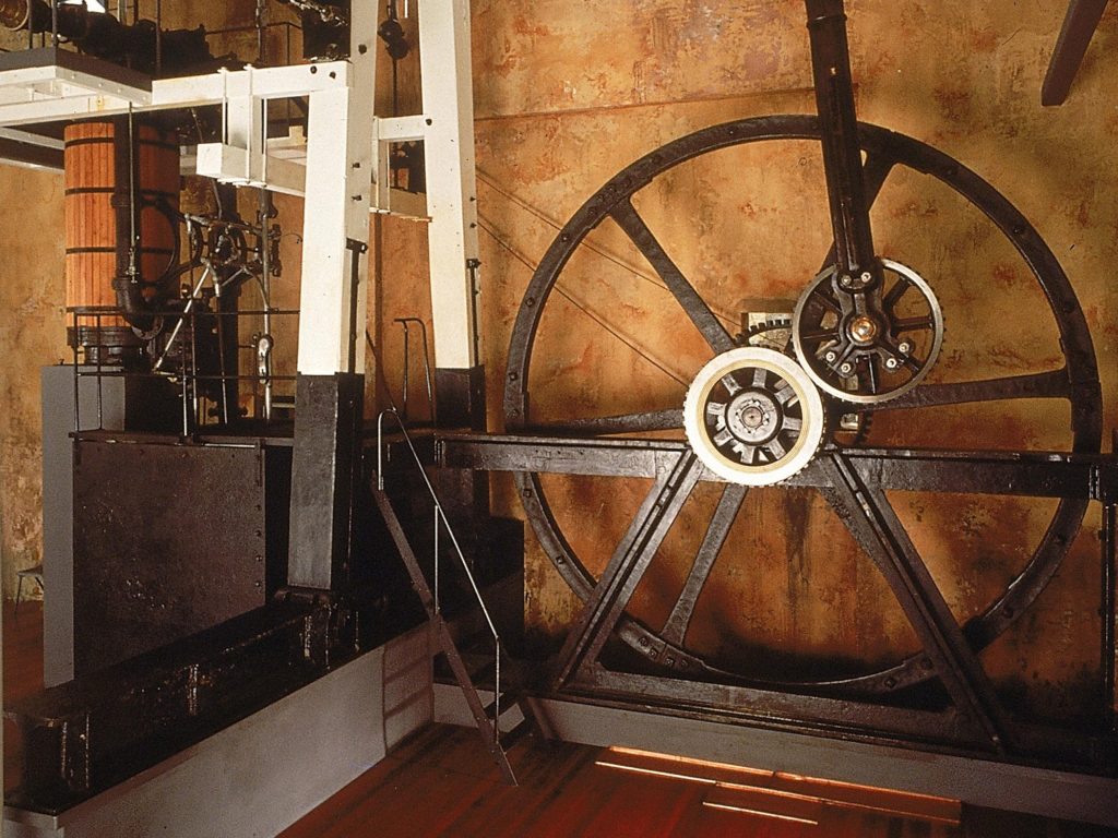 A museum display of an open steam engine made up of a large fly wheel attached by gears to a piston and steam cylinder.